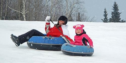 Mother and child snow tubing down a snowy hill, smiling joyfully while enjoying a fun winter adventure together.