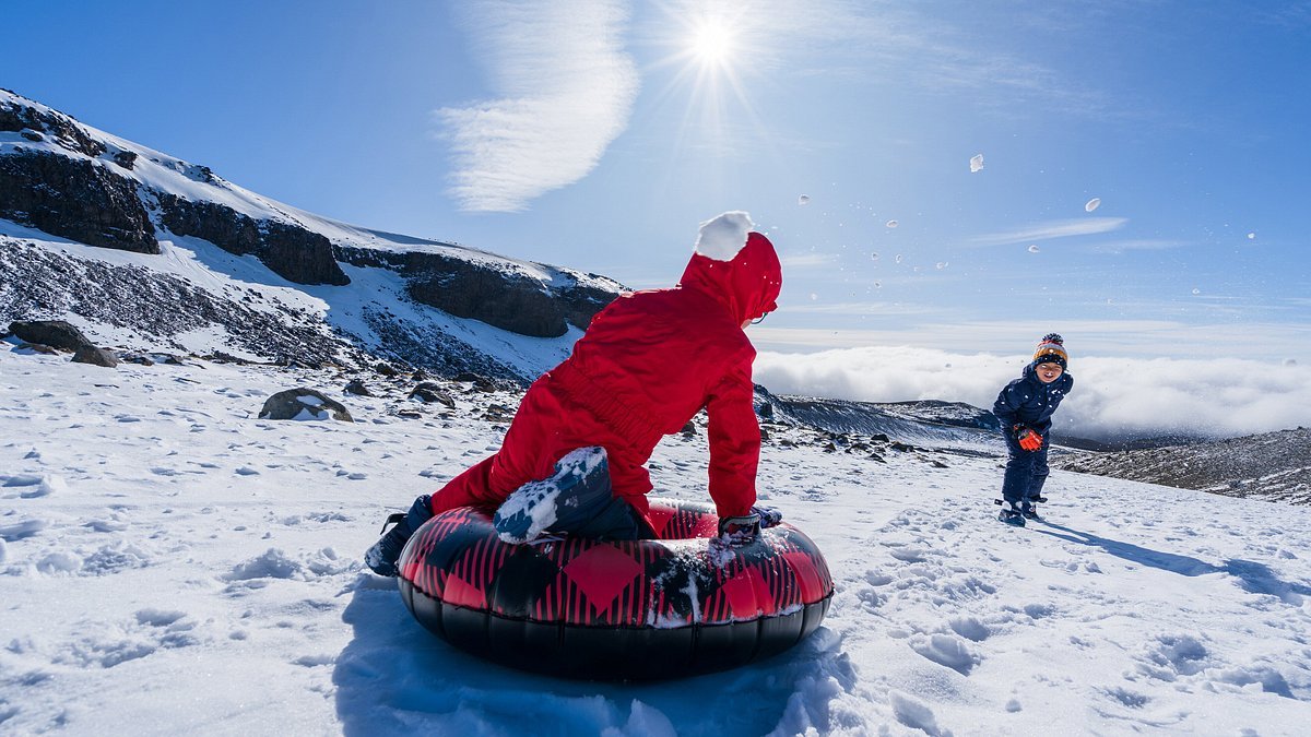 Two kids in snow gear sledding on a snow tube down a snowy hill under a sunny blue sky, enjoying winter fun.