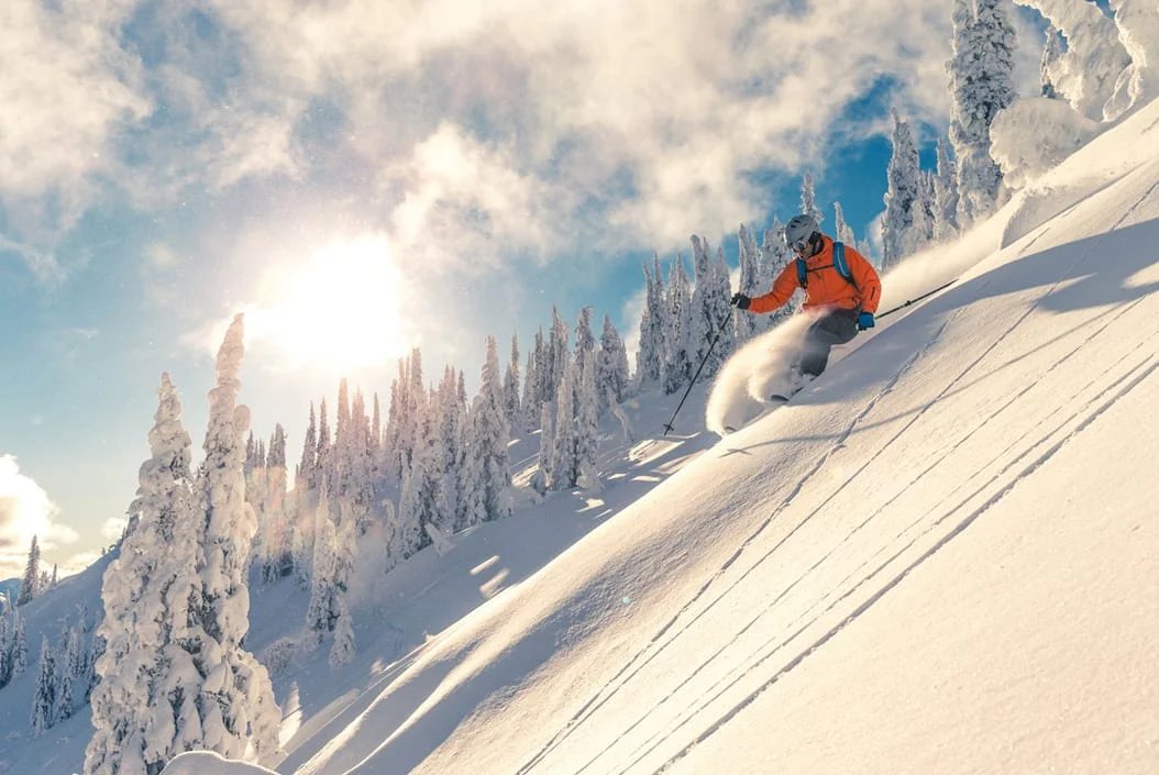Skier in bright orange gear descends a snowy mountain slope under a clear blue sky and shining sun.