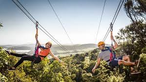 Two people ziplining over a lush forest in the Manali Valley, surrounded by breathtaking mountain scenery.