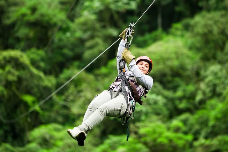 A female wearing safety gear and smiling while ziplining through the lush green forest of Manali.